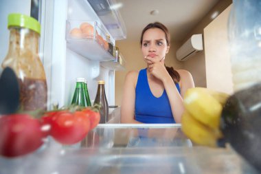 Genç kadın buzdolabını açıyor, içine bakıyor ve ne yiyeceğini seçiyor. Buzdolabı sebzelerle dolu. Buzdolabının içinden bak. Yemek, diyet, duygu, ev hayatı, zevk kavramı