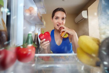 Genç kadın buzdolabını açıyor, içine bakıyor ve ne yiyeceğini seçiyor. Sosis ve sebze yemek. Buzdolabının içinden bak. Yemek, diyet, duygu, ev hayatı, zevk kavramı