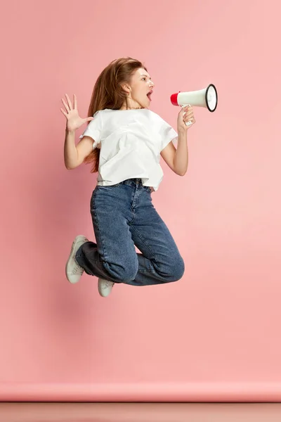 stock image Full-length portrait of young woman in casual clothes cheerfully jumping and emotionally shouting in megaphone against pink studio background. Concept of human emotions, lifestyle, youth, fashion, ad