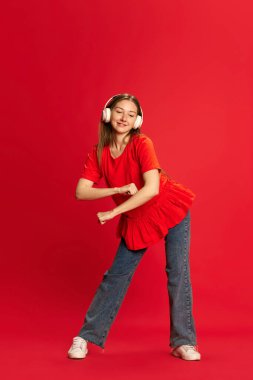 Gündelik giysiler içinde, kulaklıkla müzik dinleyen ve kırmızı stüdyo arka planında dans eden genç güzel bir kızın tam boy portresi. İnsan duyguları kavramı, yaşam tarzı, gençlik, moda, reklam