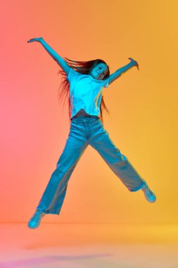 Neşe. Aktif, heyecanlı, günlük kıyafetler giyen, neon ışıklı arka planda yüksekten atlayan bir çocuk. Çocukluk kavramı, duygular, çocuk modası. Model görünümü