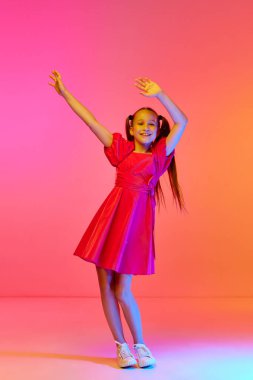 Pembe neon arka planda at kuyruğu pozu veren şenlikli elbiseli sevimli küçük bir kız. Çocukluk kavramı, duygular, çocuklar moda, dans, müzik.