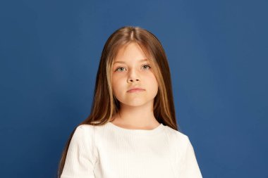 Calm look. Half-length portrait of charming little girl looking at camera over blue background. Kids emotions, fashion and beauty concept.