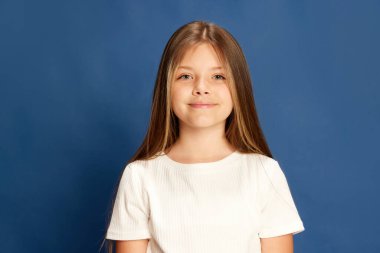 Child with light smile. Half-length portrait of charming little girl looking at camera over blue background. Kids emotions, fashion and beauty concept.