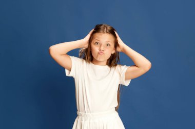 Front view grimacing little girl. Close up head shot portrait with little brown-haired girl looking at camera on blue background. Concept chilhood, happy, beauty, healthy kid. Ad