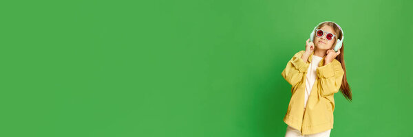 Relaxing girl with earphones. Wide photo little girl wearing stylishly, listening music over green background with smile looking at camera. Concept of technology, generation, childhood, lifestyle, ad.