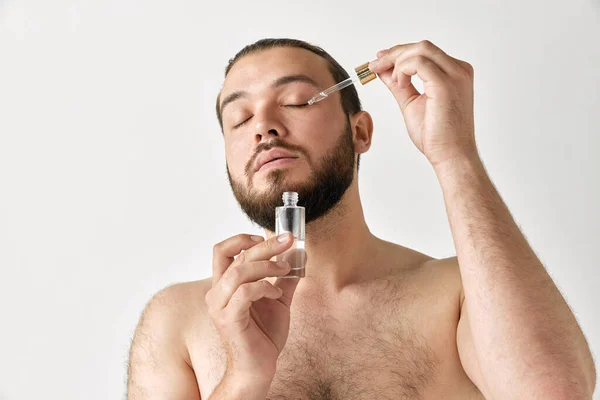 stock image Man with closed eyes enjoys application of cosmetic product from bottle he holds in hand over grey background. Concept of beauty, appearance, self care, cosmetic, procedures, health, natural, ad.