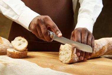 Afro-amerikalı bir adam, ahşap masada mutfak bıçağıyla taze beyaz ekmek dilimler. Sağlıklı yeme, beslenme ve el işi ürünü kavramı.