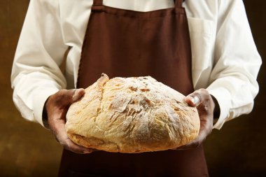 Önlüklü tanınmayan fırıncı elinde kahverengi arka planda bir somun ekmek tutuyor. Yemek, sağlık, gelenek, fırın ve yemek yapma kavramı. Ad