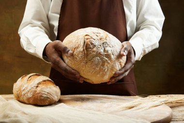 Kahverengi önlüklü tanınmayan Afrikalı fırıncı elinde yuvarlak ekmek, yanında da kahverengi arka planda uzun bir somun var. Yemek, sağlık, gelenek, fırın ve yemek yapma kavramı. Ad