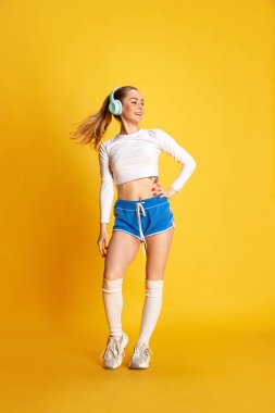 Genç, gülümseyen, kulaklıklı, sarı arka planda gözlerini çevirmiş bir kadının dikey fotoğrafı. Spor anlayışı, hobi, fitness, sağlıklı yaşam tarzı, reklam