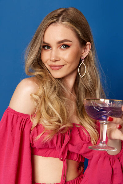 Portrait of charming girl dressed bright and stylishly holding cocktail and looking at camera over blue studio background. Concept of fashion, party, beauty, human emotions