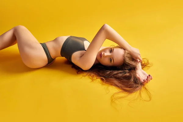 Photo of young woman with fit body wearing khaki underwear posing on floor and looking at camera over yellow background. Concept of natural beauty, body and skin care, health, wellness, femininity. Ad