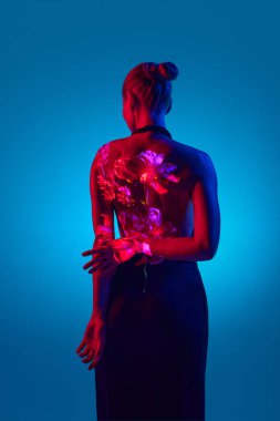 Arkadan bak. Dijital neon projektörlü güzel bir kadın arkadaki mavi renkte çiçekleri gösteriyor. Dijital sanat, moda, fütürizm, vücut, kavram