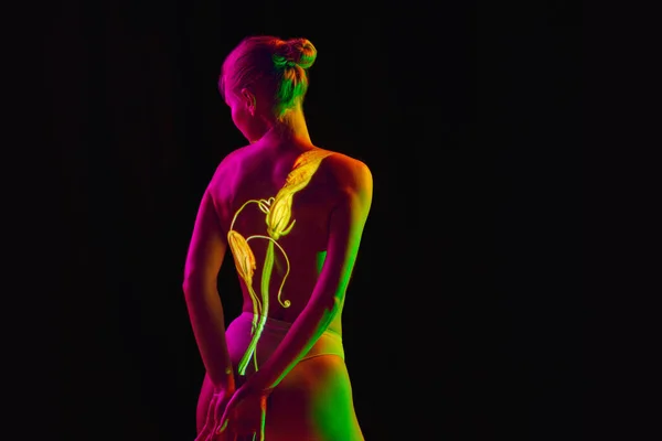 Back view. Image of naked young slim woman posing in neon light with flower reflection isolated on dark mode background. Copy space for ad Concept of emotions, beauty, expression, youth, aspiration.