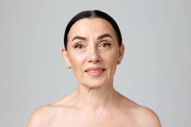 Çıplak omuzlu güzel, olgun, esmer kadın gri arka planda kameraya bakıyor. Sağlık hizmetleri. Moda, güzellik, kaplıca, kozmetoloji, cilt bakımı, kadınlık kavramı.