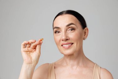 Gri arka planda kozmetik yağ kapsülünü tutan bej iç çamaşırlı muhteşem esmer kadının portresi. Moda, güzellik, kaplıca, kozmetoloji, saç bakımı ve sağlık konsepti.