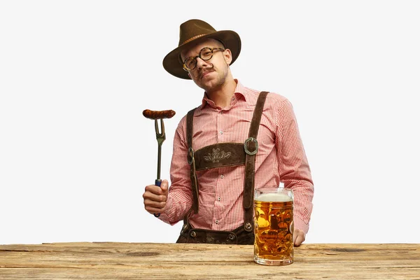 Bavarian mustache man in hat wearing traditional fest outfit holding hot fried sausage and looking at huge glass of beer. Concept of Oktoberfest, traditions, drinks and food. Copy space for ad