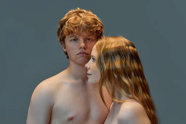 Shirtless redhead man looking at camera, standing near his redhead girlfriend who stands sideways looking away against gray background. Concept of natural beauty, love, relationships, fashion. Ad