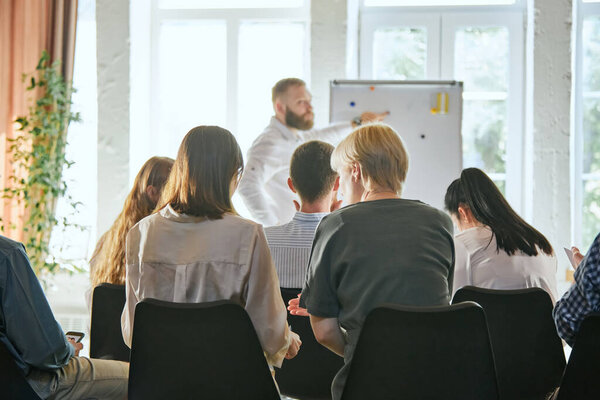 Мужчина выступает с докладом на деловой встрече. Задний вид аудитории в конференц-зале. Женщины обсуждают информацию. Концепция конференции, обучения, планирования, обучения, коучинга, бизнеса.