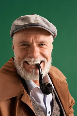 Kendinden emin, olgun bir adamın fotoğrafı. Klasik şapka takan dedektif gibi görünüyor. Ağzında piposu olan çizgili takım elbise ve yeşil arkaplan. Moda kavramı, duygular, retro