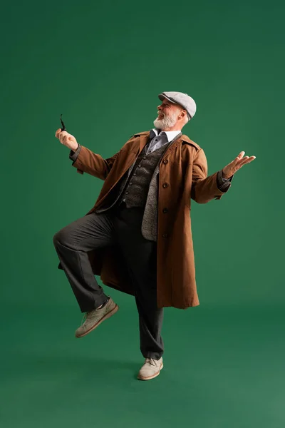 Full lenght photo. Emotional mature man with mustache, dressed in vintage style, looks like detective, laughing while dancing. Concept of fashion, emotions, vintage, retro and ad