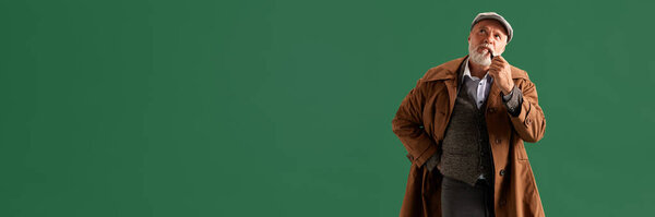 Photo of confident, positive mature man, looks like detective wearing vintage hat, striped suit with pipe in mouth against green color background. Concept of fashion, emotions, retro. Ad. Banner.