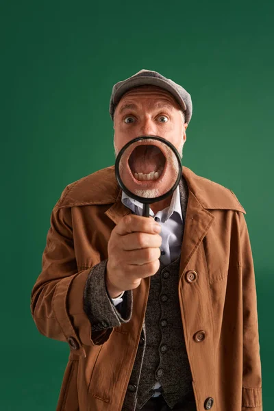 Half-length portrait of funny man, detective wearing vintage hat, suit with magnifying glass near face throught which can see his open mouth. Concept of fashion, emotions, retro. Ad.
