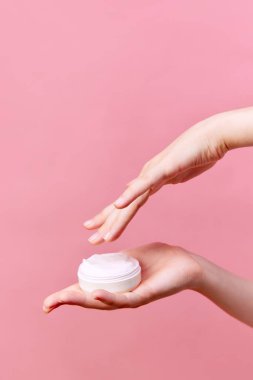 Ellerinde krema kavanozu tutan kadın ellerinin kesilmiş fotoğrafı. Kozmetik ürünleri kullanarak izole edilmiş pastel pembe arka plan. Vücut ve cilt bakımı, kaplıca, cilt bakımı. İlan için alanı kopyala