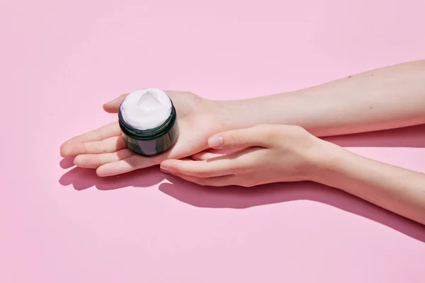 Cropped top view of female hands holding black jar of cream, night cream in hands. Using cosmetics product isolated pastel pink background.Concept of body and skin care, spa, dermatology, anti-aging
