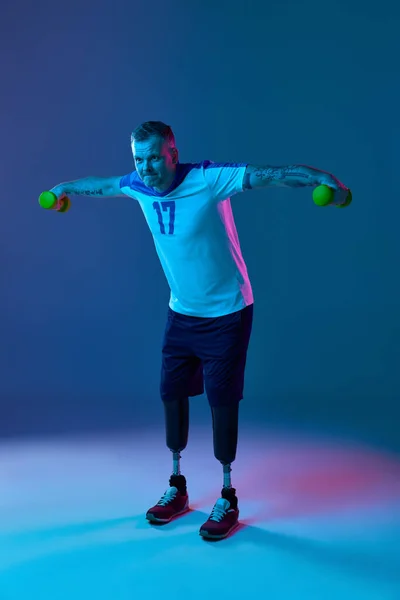 Young muscular man with prosthetic legs doing exercises in gym with dumbbells in neon light. Motivation poster. Concept of sport for people with disabilities, medical, health care.