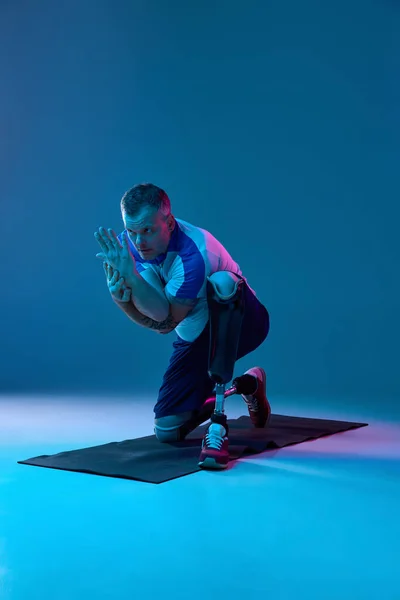 Confident man in sport uniform prosthetic legs doing exercises for hands in neon light on floor. Motivation poster. Concept of sport for people with disabilities, medical, health care.