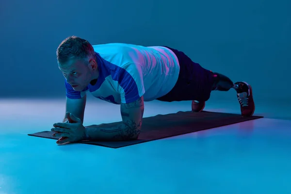 Confident man in sport uniform prosthetic legs doing plank exercise in horizontal position in neon light on floor. Motivation poster. Concept of sport for people with disabilities, medical, health.