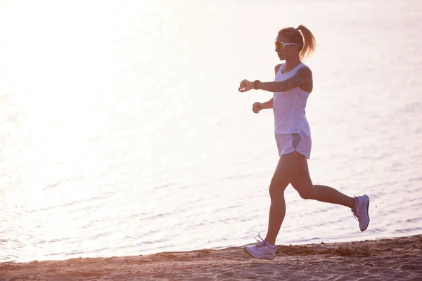 Young fit woman training, running with sports tracker at beach near river on sunrise in motion. Morning fitness exercise on seaside. Concept of sport, recreation, healthy lifestyle, hobby
