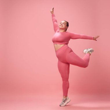 Uzun boylu, mutlu, genç, kilolu, aktif, pembe elbiseli bir kadın ve atölyenin arka planında izole edilmiş bacaklarla trenleri ısıtan taytlar. Egzersiz, spor, motivasyon, vücut pozitifliği kavramı