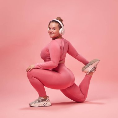 Pembe bluzlu ve bacaklı tombul bir kadının tam vücut portresi. Stüdyo arka planında müzik eşliğinde bacakları eğitiyor. Spor anlayışı, spor anlayışı, sağlıklı yaşam tarzı, motivasyon