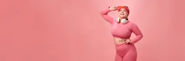 ピンクの上の若い魅力的なプラスサイズのアクティブな女性の肖像画と 隔離されたスタジオの背景を指すヘッドフォンで脚 ワークアウト スポーツ モチベーションボディのポジティブさの概念 バナー — ストック写真