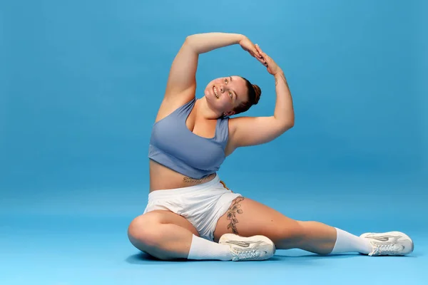 Self-care and well being. Young overweight woman stretching in sportswear against blue studio background. Motivation. Concept of sport, body-positivity, weight loss, body and health care