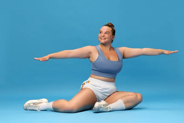 Self-care and well being. Young overweight woman stretching on floor in sportswear against blue studio background. Motivation. Concept of sport, body-positivity, weight loss, body and health care