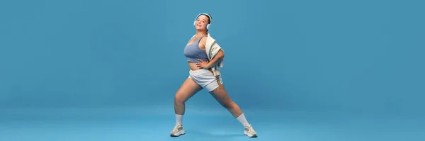 Self-care and well being. Young smiling plus-size woman doing exercises lunges in sportswear against blue studio background. Motivation. Concept of sport, body-positivity, weight loss. Banner