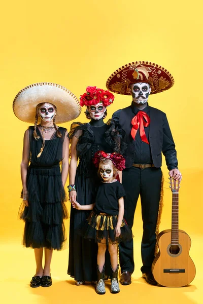 stock image Frightening family with face art of zombies celebrating Halloween in costume over bright yellow studio background. El Dia de Muertos. day of the dead. Concept of autumn, holidays, horror, body art. Ad