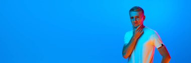 Pankart. Genç bir adamın portresi, mavi stüdyo arka planındaki öğrenci neon ışığı, filtre. Yakışıklı, dalgın erkek manken gündelik tarzda. İnsan duyguları kavramı, yüz ifadesi, gençlik, satış, reklam.