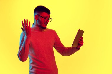 Sakallı genç adam tablet tutuyor ve sarı zemin üzerinde kırmızı ışık ve filtre ile şaşırmış görünüyor. Sabah toplantısı. İnsan duyguları kavramı, yüz ifadesi, online eğitim, iş. Ad