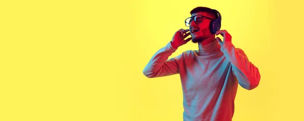 Handsome young guy in headphones listening music and sing along against on yellow background in red color light, filter. Concept of human emotions, leisure, relax. Banner. Poster. Copy space for ad.