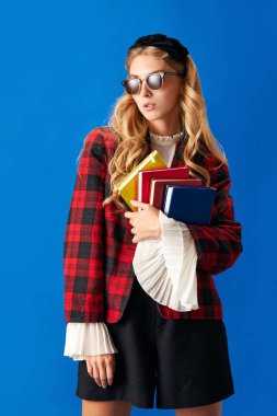 Genç bir kadının portresi, modaya uygun giyinmiş, güneş gözlüklü, elinde kitaplarla arka planda mavi bir stüdyo. Okula dönüş kavramı, eğitim, öğrenci yaşam tarzı, ödev. Ad