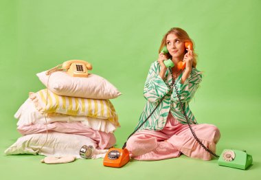 Uyandırma servisi. Pijamalı tatmin olmamış genç bir kadının tam boy portresi pastel yeşil arka planda izole edilmiş iki retro telefonla konuştu. Benim için uyanma kavramı, sağlıklı uyku, iş ve dinlenme, uyku zamanı..