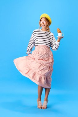 Pembe güneş gözlüklü ve bereli mutlu bayan portresi mavi arka planda bir fincan kahveyle lezzetli kruvasan yiyor. Yemek, mutfak, moda, tarz, yolculuk, macera kavramı. Ad