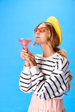 Şık giyinmiş, güneş gözlüklü ve sarı bereli bayan portresi mavi arka planda. Yiyecek ve içecek kavramı, alkol, mutfak, moda, tarz, macera. Ad