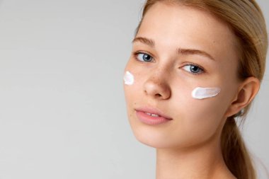 Gri stüdyo arka planına izole edilmiş lekesiz, bakımlı yüz kremi olan güzel bir kızın yakın plan portresi. Cilt bakımı kavramı, doğal güzellik, kozmetik ve kozmetik, spa