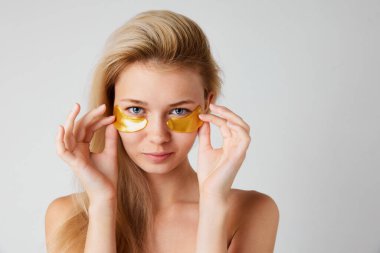 Gri stüdyo arka planında izole edilmiş göz altı bantları kullanan iyi korunmuş ciltli genç bir kız. İçecek bir şeyler. Cilt bakımı kavramı, doğal güzellik, kozmetik ve kozmetik, spa